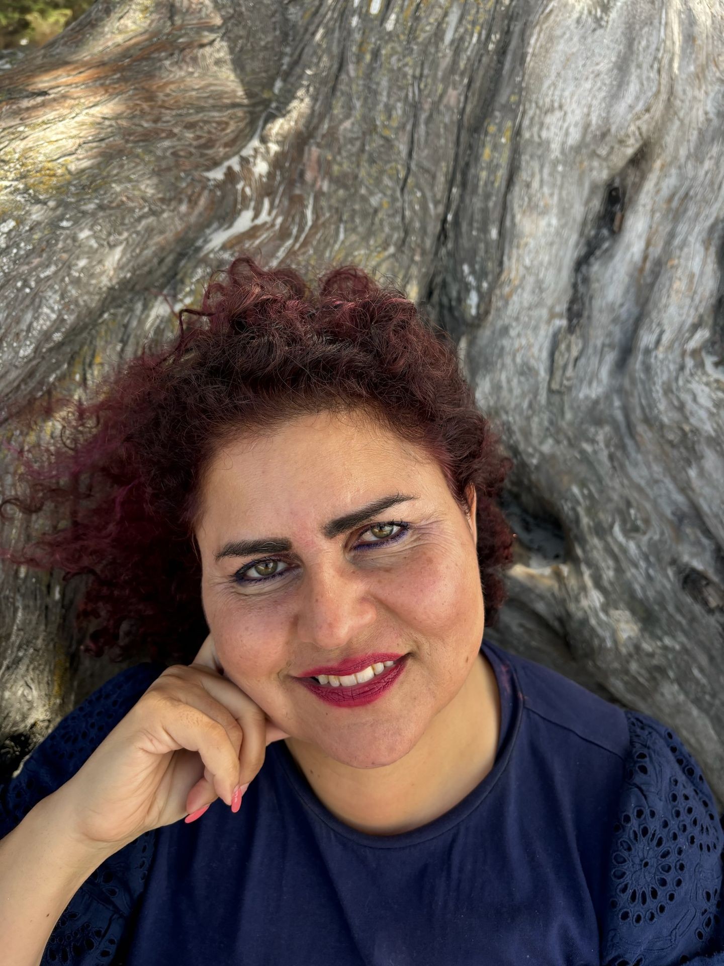 Smiling woman with curly hair and blue top, sitting in front of a textured tree trunk.